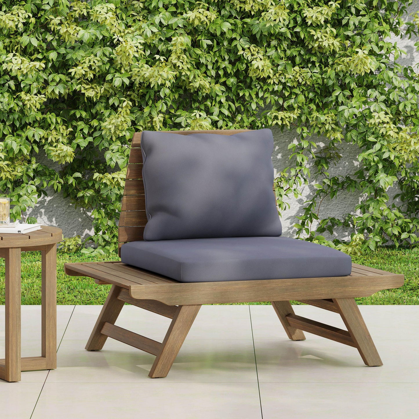 The Sedona Outdoor Club Chair with a slatted design and gray cushions sits on a tiled patio beside a small side table, framed by a lush green vine-covered wall.