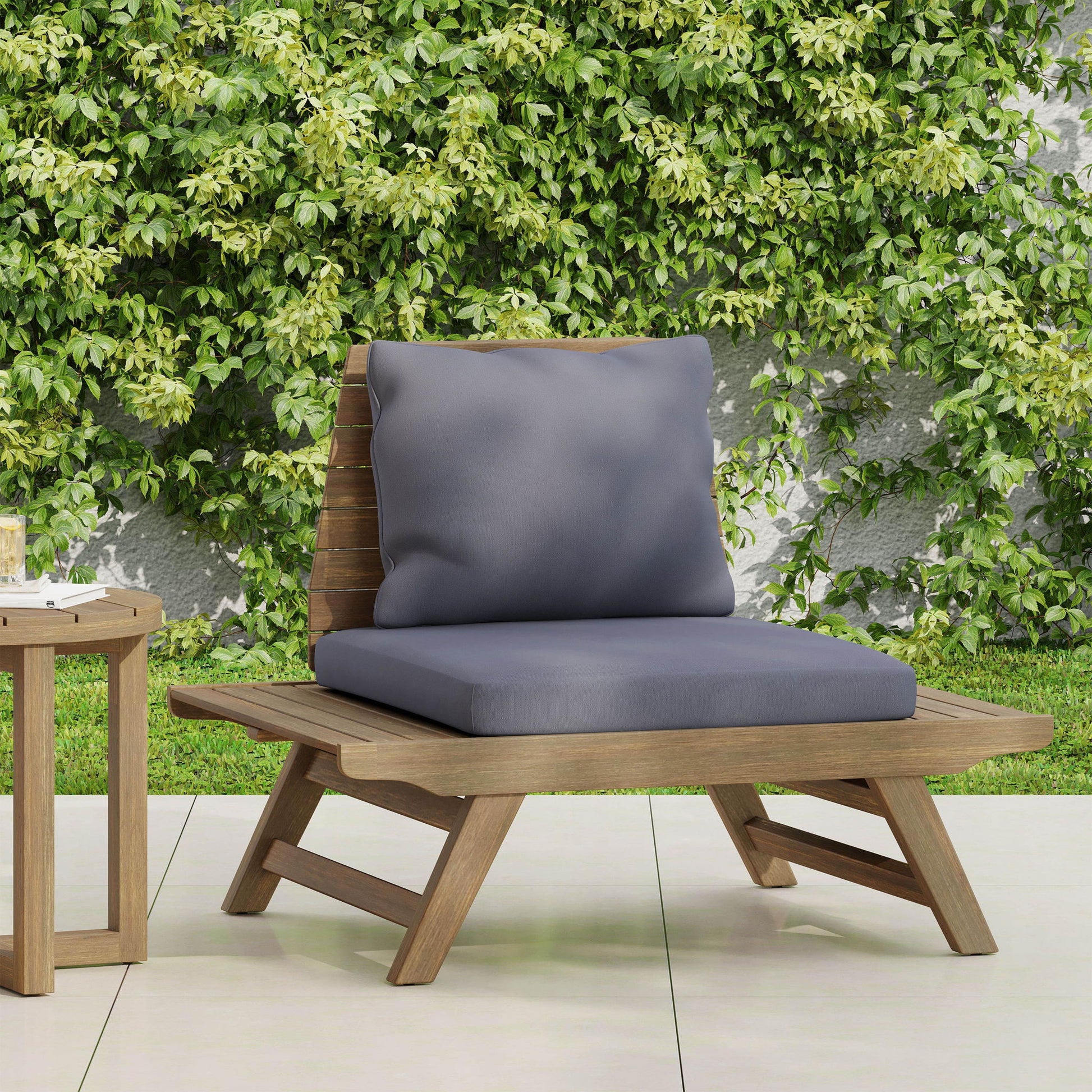 The Sedona Outdoor Club Chair with a slatted design and gray cushions sits on a tiled patio beside a small side table, framed by a lush green vine-covered wall.