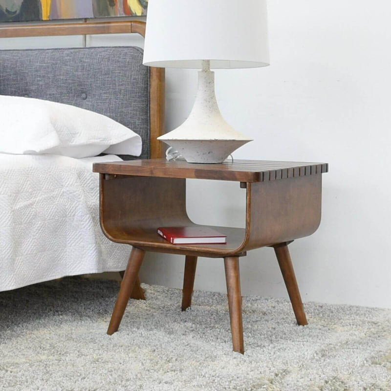 The Alice - Mid-Century Modern Table with angled legs sits on a gray carpet by the bed, holding a white lamp and a red book on top, with an open shelf beneath the tabletop.