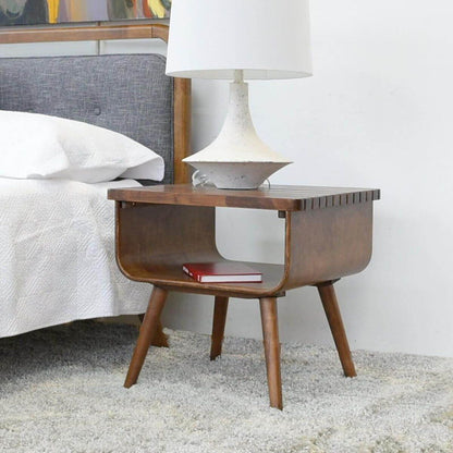 The Alice - Mid-Century Modern Table with angled legs sits on a gray carpet by the bed, holding a white lamp and a red book on top, with an open shelf beneath the tabletop.