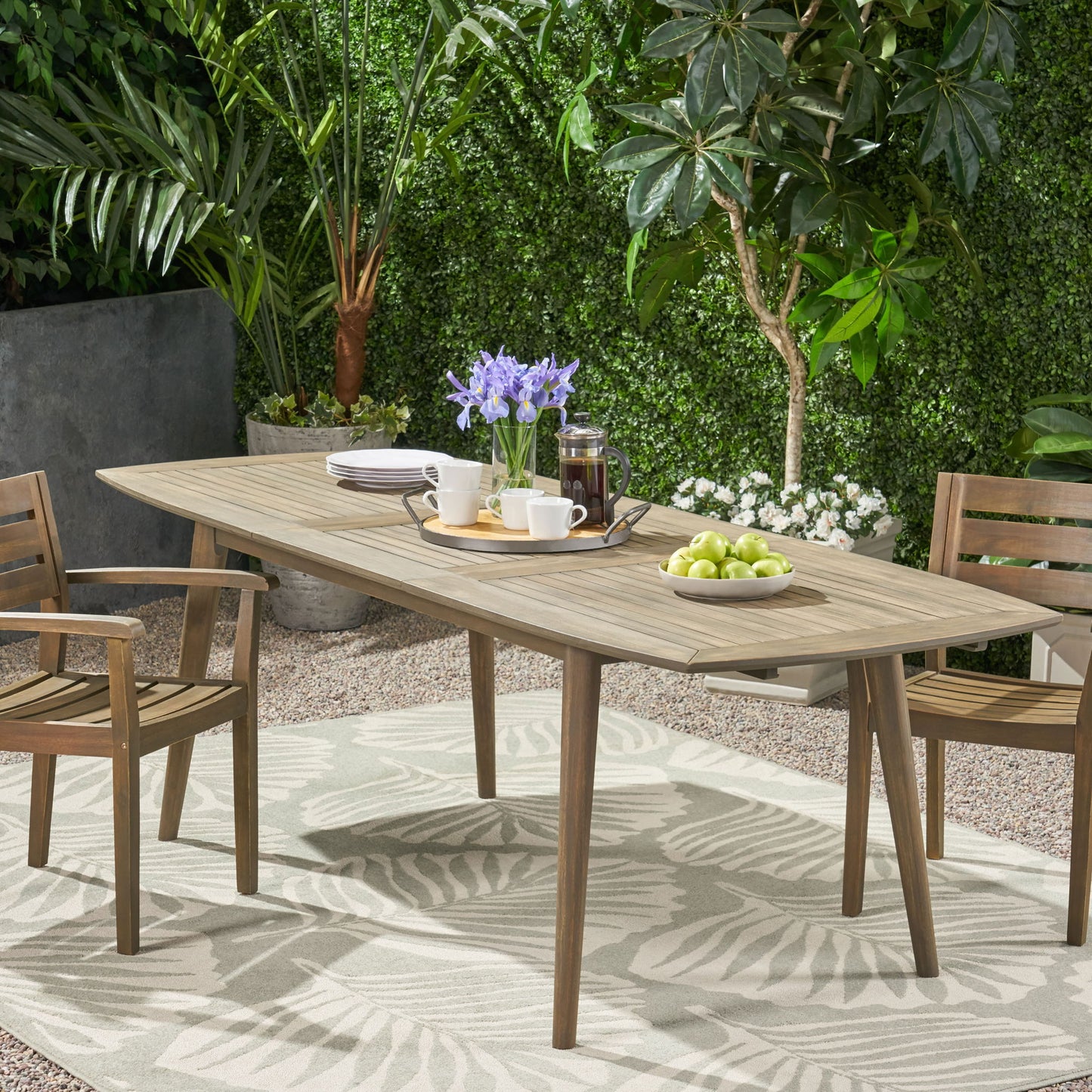 The Placidity Outdoor Expandable Dining Table with slatted tabletop and tapered legs is paired with two chairs on a leafy rug. A tray with tea, cups, plates, and green apples sits atop, surrounded by lush greenery for a serene patio setting.
