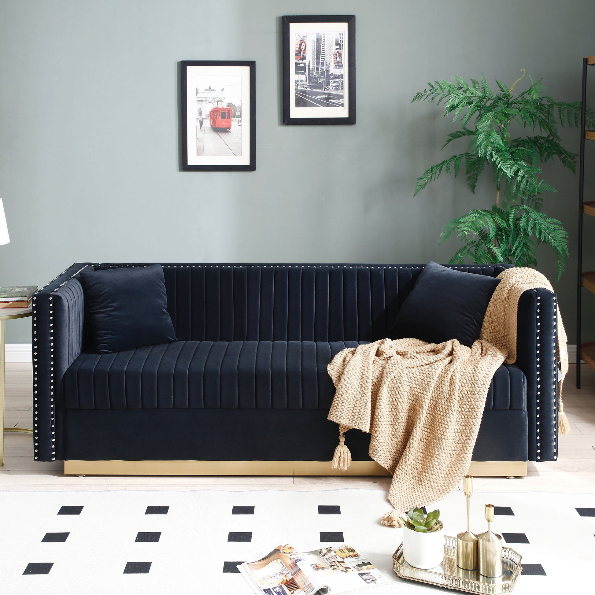 A modern living room features the Contemporary Vertical Channel Tufted Velvet Sofa Loveseat 2-Piece Set with 4 pillows in dark blue, a beige knit throw, potted plant, 2 framed cityscape photos on a gray wall, gold tray on geometric rug, and corner bookshelf.