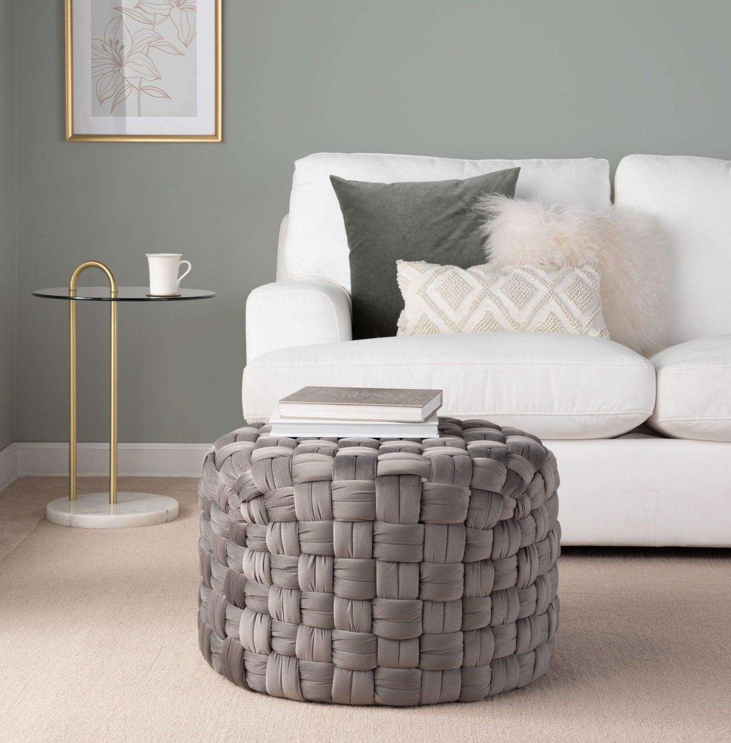 A modern living room with a white sofa, textured pillows, a round glass side table with a gold base, and the Braided - Versatile Round Ottoman in gray topped with books on a beige carpet.