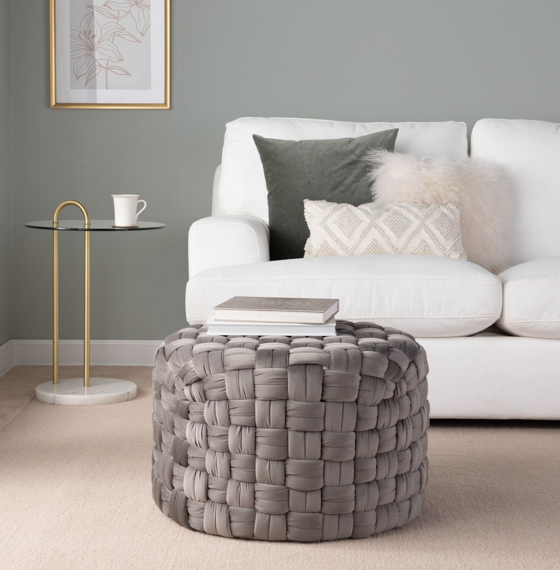 A modern living room with a white sofa, textured pillows, a round glass side table with a gold base, and the Braided - Versatile Round Ottoman in gray topped with books on a beige carpet.