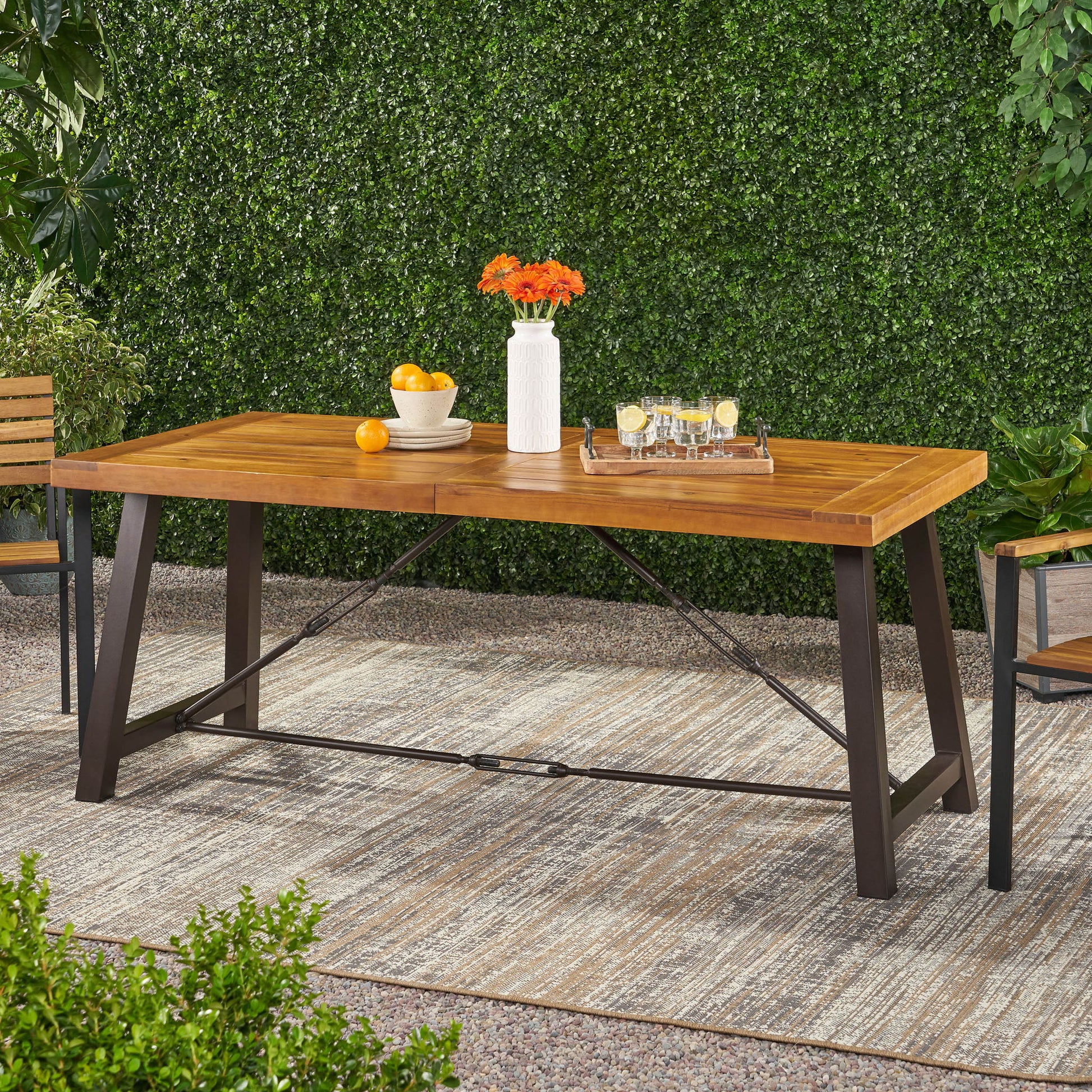 The Outdoor Dining Table With Slat Wooden Panel And Turnbuckle Accents - Teak, featuring black metal legs, stands on a patio rug with a vase of orange flowers, a bowl of oranges, and drinks. Lush green hedges and plants fill the background.