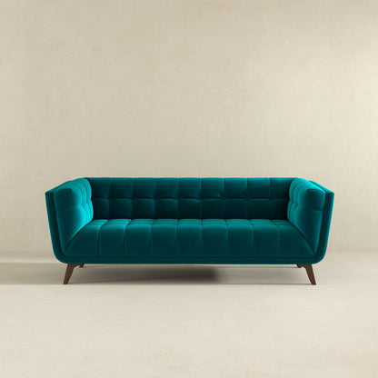 The Addison Mid-Century Modern Design Tufted Sofa, in teal velvet with tufted cushions and angled wooden legs, is set against a plain beige wall and a light floor.