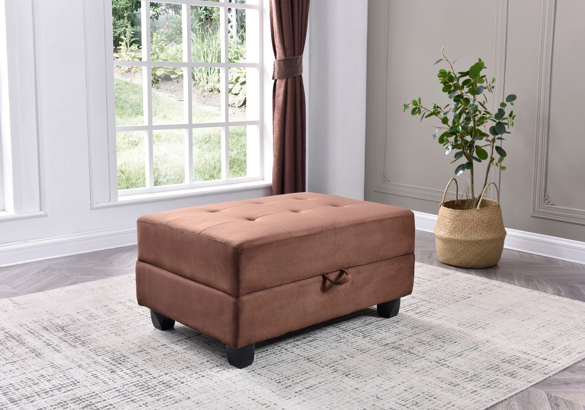 The Stylish Storage Contemporary Ottoman with a handle sits on a light patterned rug in a living room with light gray walls, a large curtained window, and a potted plant in a woven basket nearby.