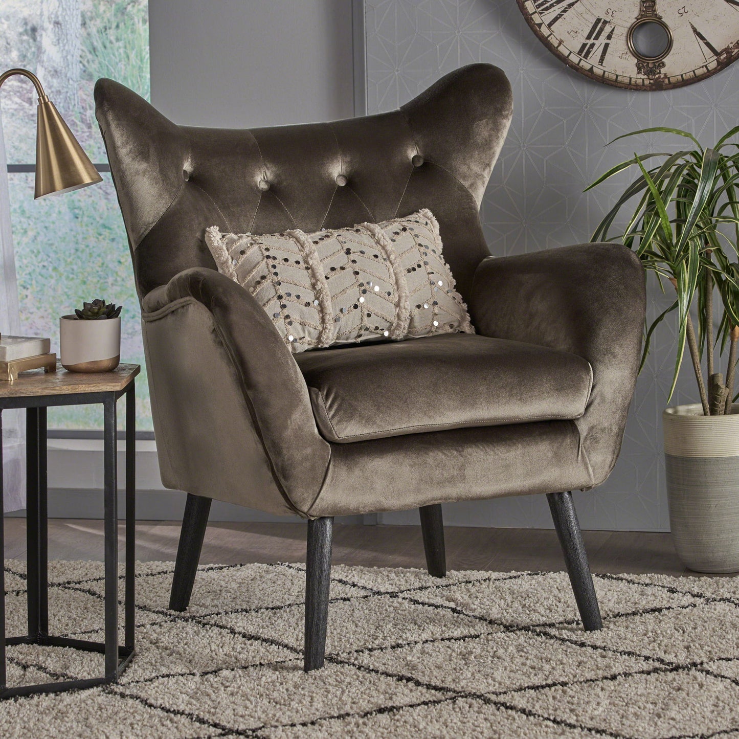 The Alyssa Mid Century Upholstered Arm Chair with wingback design, featuring plush dark gray velvet and button-tufted backrest, stands on wooden legs beside a small table, potted plant, and wall clock in a modern living room.