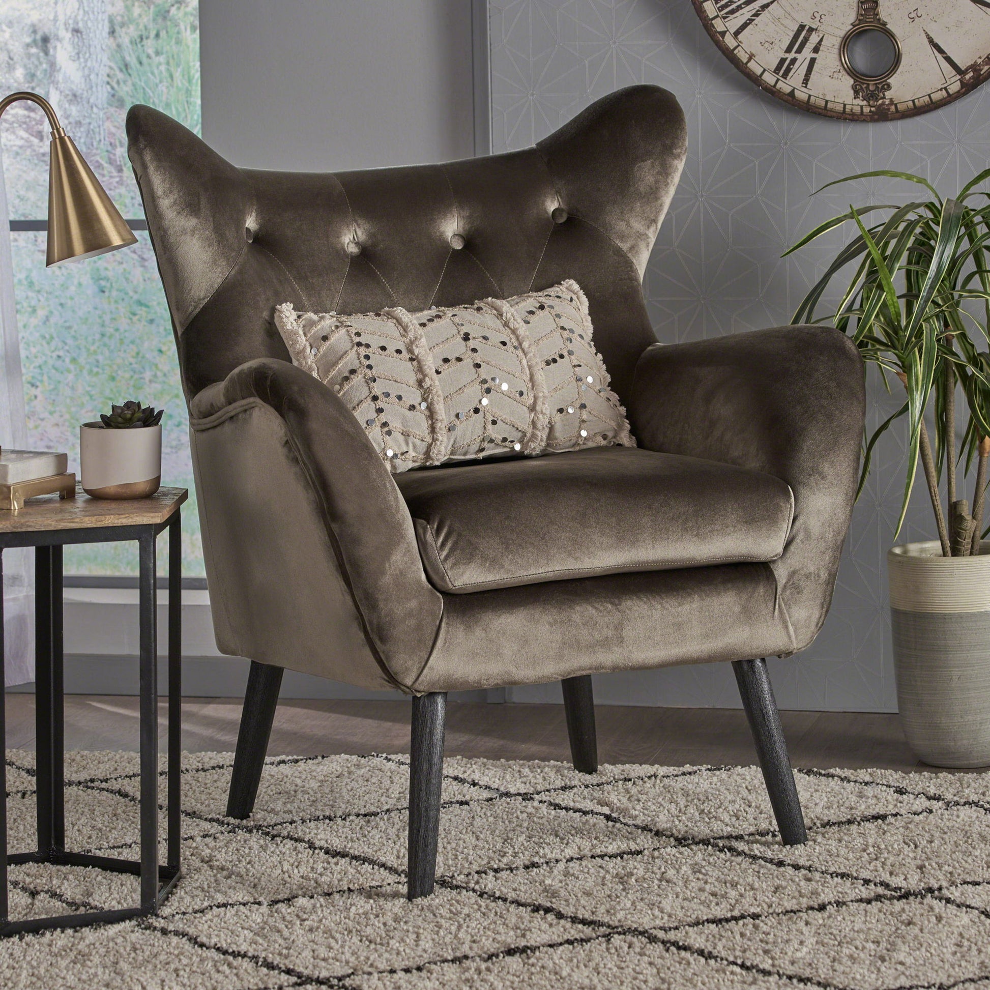 The Alyssa Mid Century Upholstered Arm Chair with wingback design, featuring plush dark gray velvet and button-tufted backrest, stands on wooden legs beside a small table, potted plant, and wall clock in a modern living room.
