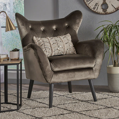 The Alyssa Mid Century Upholstered Arm Chair with wingback design, featuring plush dark gray velvet and button-tufted backrest, stands on wooden legs beside a small table, potted plant, and wall clock in a modern living room.