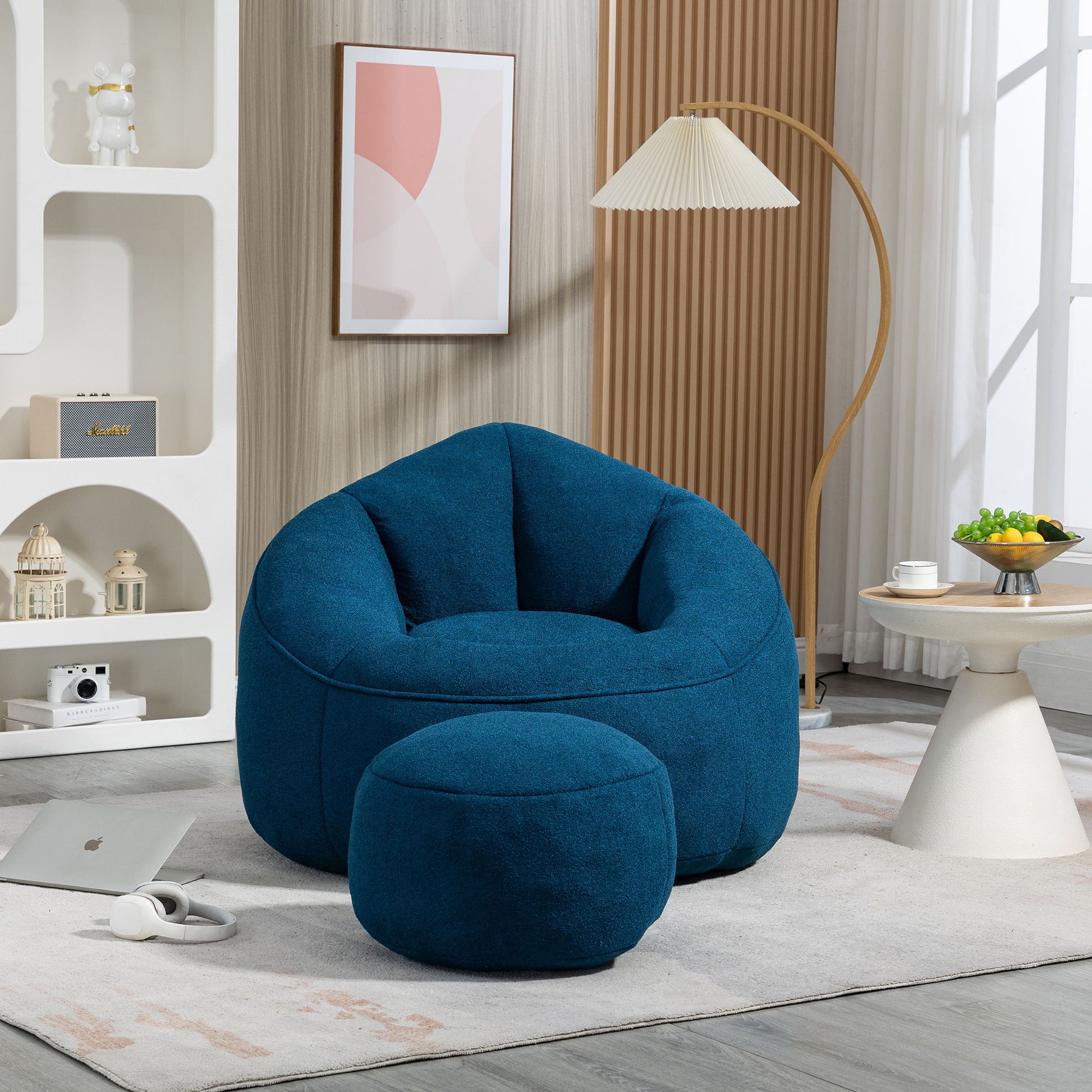 A cozy living room features the Bedding Bean Bag Sofa Chair High Pressure Foam with Footrest on a light rug, next to a round table, floor lamp, abstract wall art, and decor shelves. Sunlight pours through a large window.
