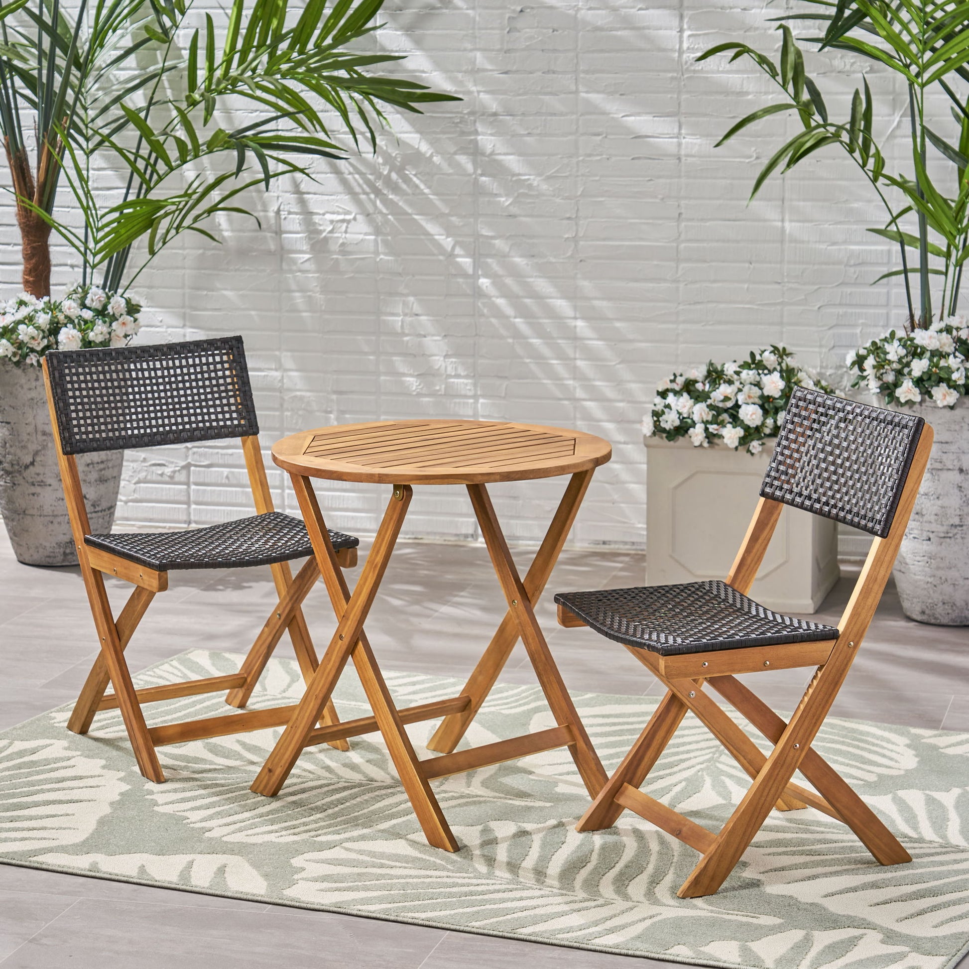 The Evergreen Outdoor Dining Set, featuring a dining table and two chairs, is arranged on a patterned rug with potted plants, white flowers, and a white brick wall in the background.