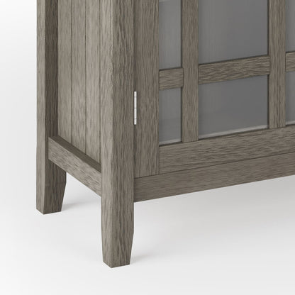 Close-up of the bottom corner of the Bedford - Storage Media Cabinet, featuring gray wood, glass panel doors, and simple straight legs.