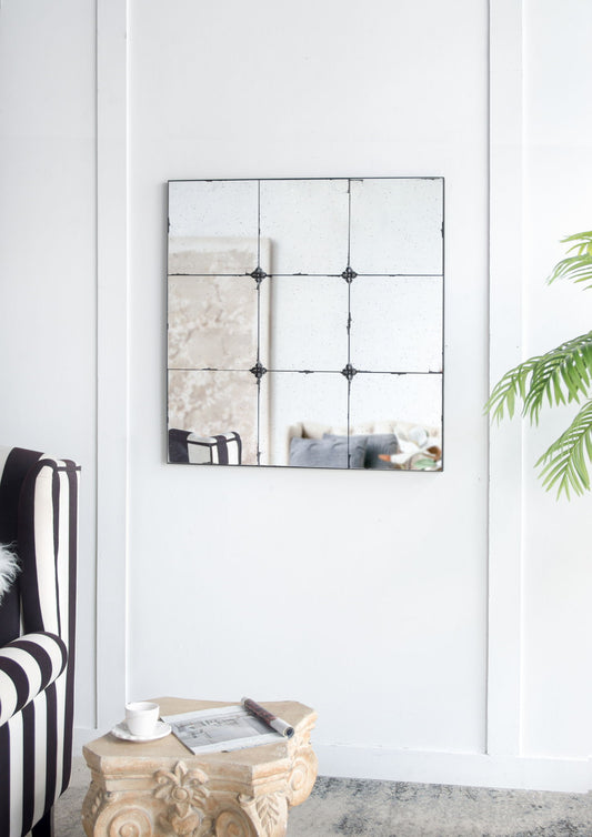 A modern living room includes the Traditional Decor Style Antique Glass Panel Mirror, a striped black-and-white armchair, a carved stone table with a cup and magazine, and a leafy green plant against white walls.