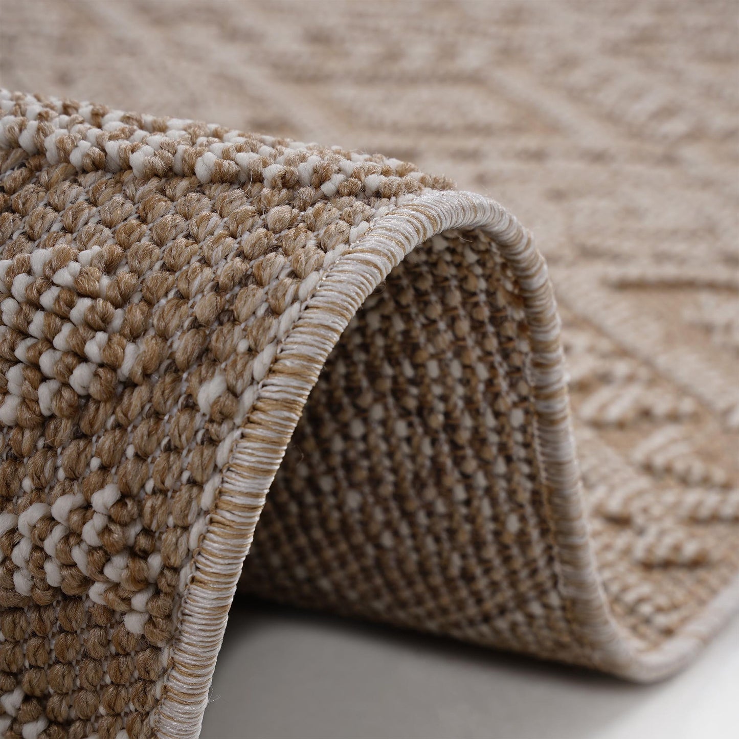 Close-up of the Guros - Traditional Indoor / Outdoor Area Rug in beige and cream, with a curved edge lifted to reveal its detailed textured patterns and stitched border.