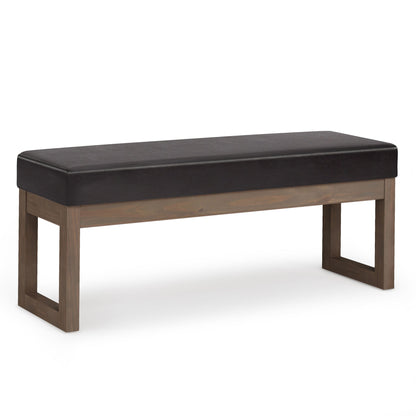 The Milltown Upholstered Ottoman Bench features a dark cushioned seat and a sturdy, rectangular wooden frame in a natural finish with open square legs at each end.