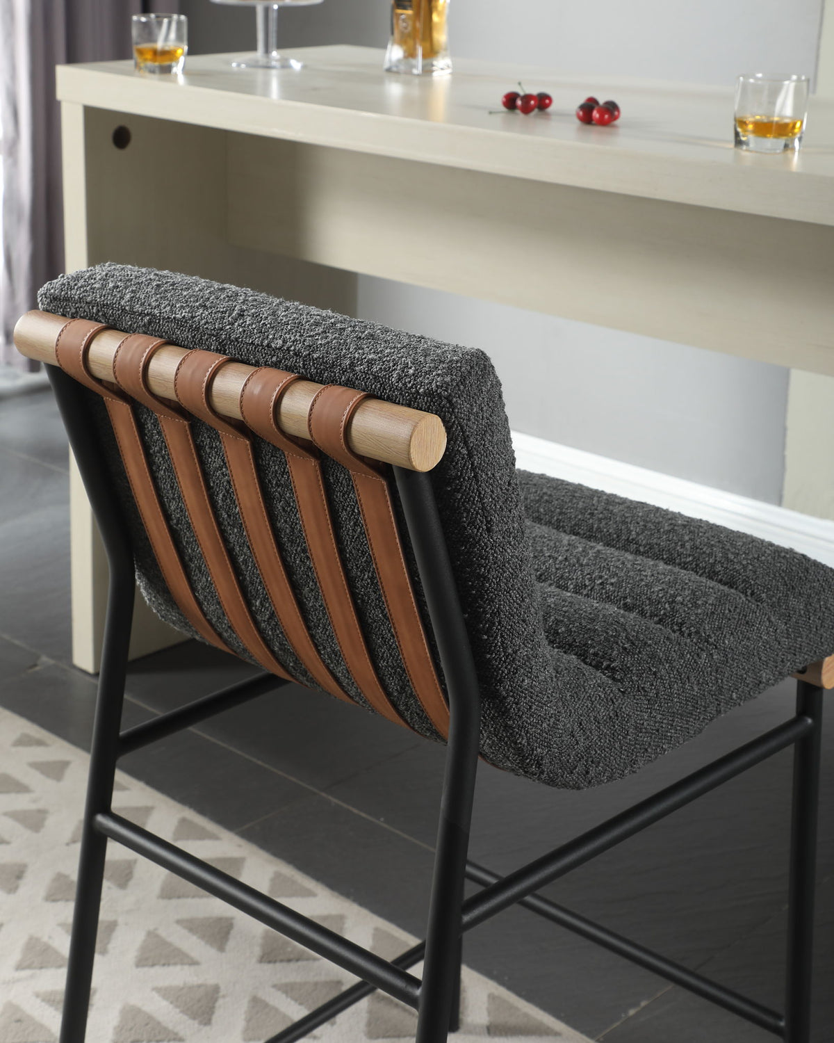 The Lahni 17" Boucle Fabric Counter Height Chair, featuring a textured gray cushion, leather strap accents, and black metal legs, stands beside a light bar topped with drinks and scattered cherries.