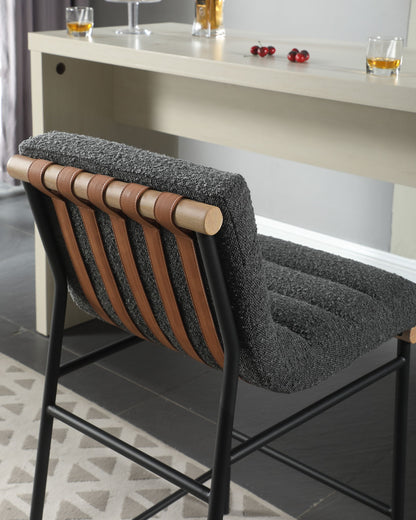 The Lahni 17" Boucle Fabric Counter Height Chair, featuring a textured gray cushion, leather strap accents, and black metal legs, stands beside a light bar topped with drinks and scattered cherries.