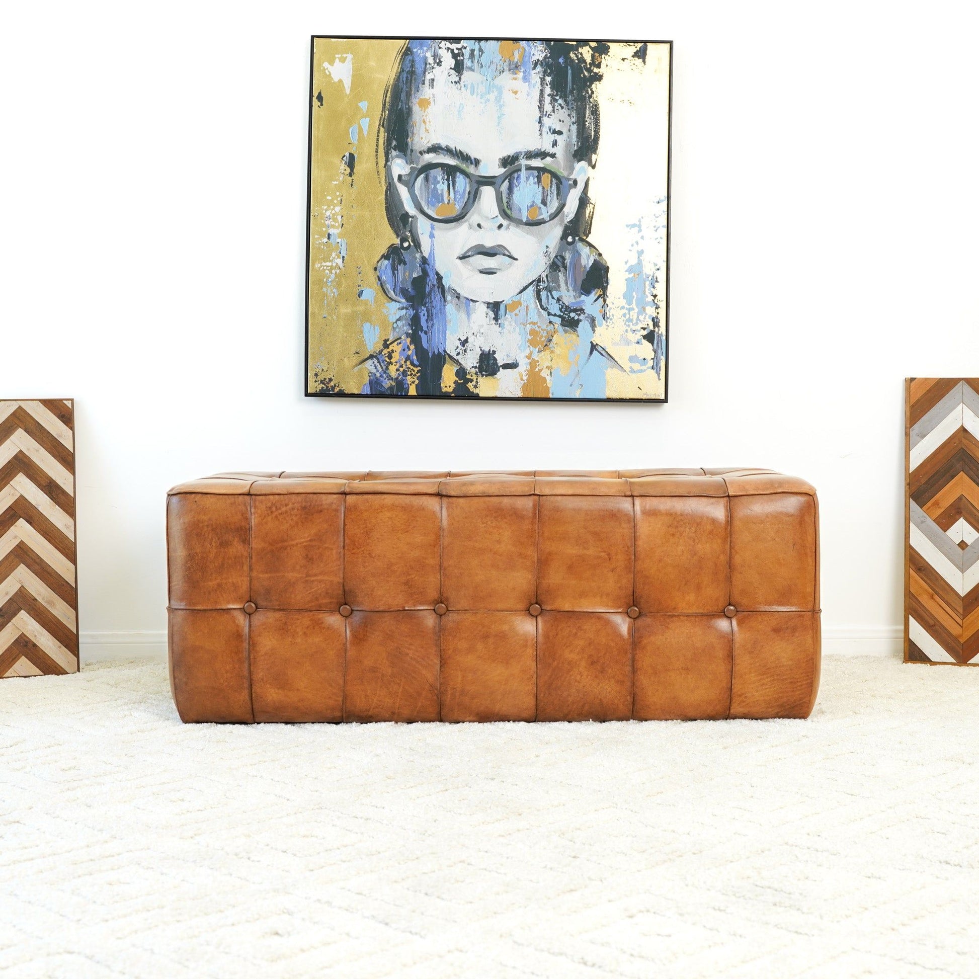 The Benjiro - Bench - Tan sits on a cream textured rug. Above it hangs a colorful portrait of a woman in sunglasses, while two chevron-patterned geometric artworks lean against the white wall on either side.