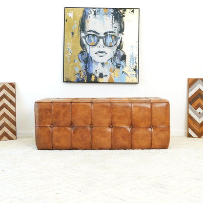 The Benjiro - Bench - Tan sits on a cream textured rug. Above it hangs a colorful portrait of a woman in sunglasses, while two chevron-patterned geometric artworks lean against the white wall on either side.