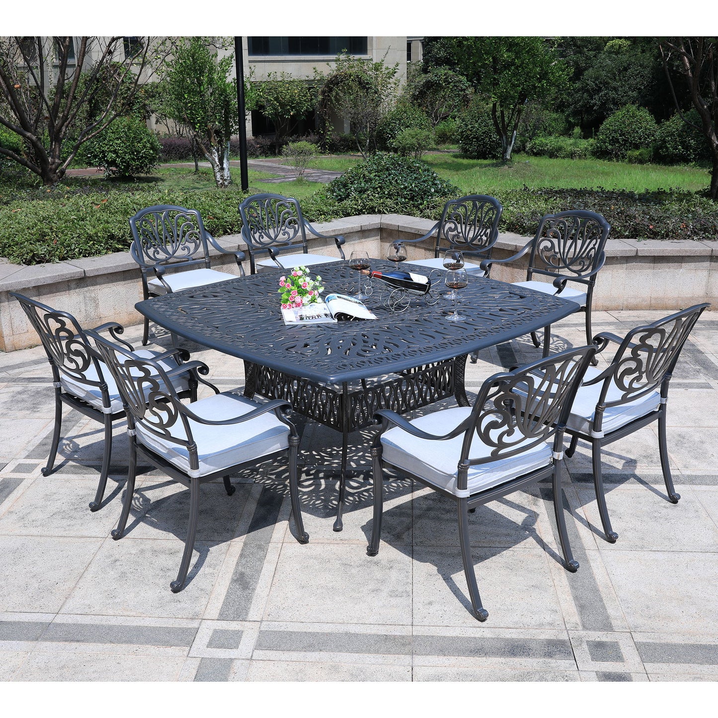 The Square 8 Person 64" Long Aluminum Dining Set With Cushions features a black ornate table and eight cushioned chairs, arranged in a garden with greenery and stone pavement. The table is set with place settings and a flower vase.