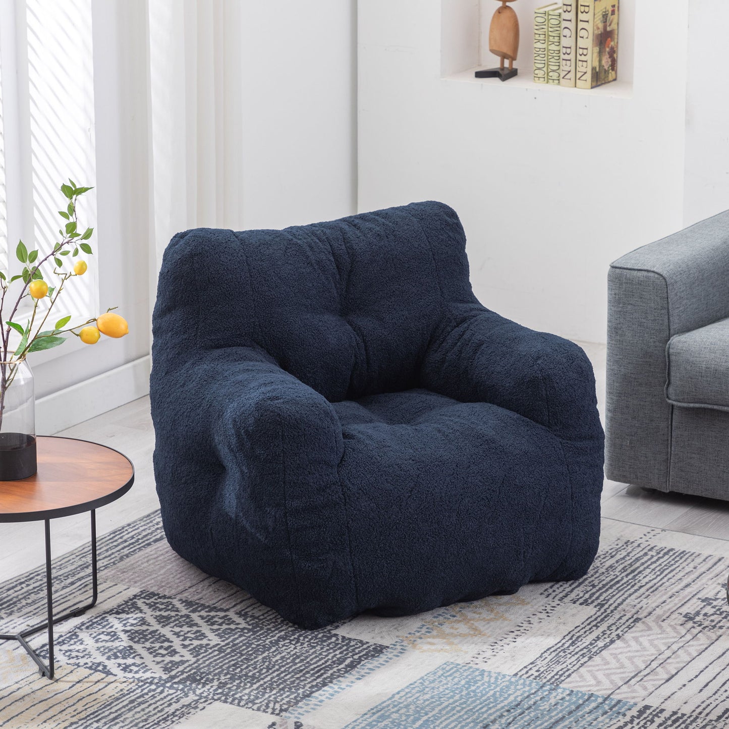 A Soft Teddy Fabric Tufted Foam Bean Bag Chair sits in a bright living room next to a small round table with a vase of yellow lemons, near a gray sofa and books on built-in shelves.