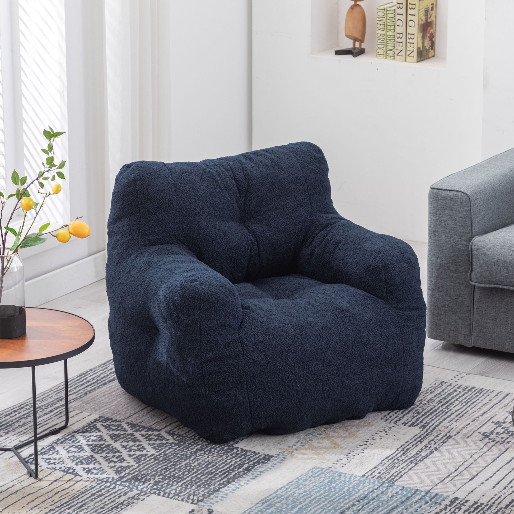 A Soft Teddy Fabric Tufted Foam Bean Bag Chair sits in a bright living room next to a small round table with a vase of yellow lemons, near a gray sofa and books on built-in shelves.