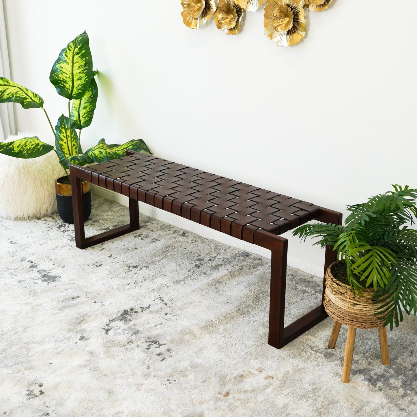 The Christina Leather Bench in Cognac, Dark Brown, and Tan sits on a light patterned rug, surrounded by green potted plants and accented by gold floral wall art on a white wall.
