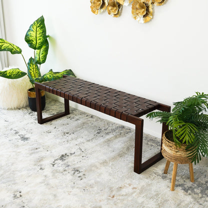 The Christina Leather Bench in Cognac, Dark Brown, and Tan sits on a light patterned rug, surrounded by green potted plants and accented by gold floral wall art on a white wall.