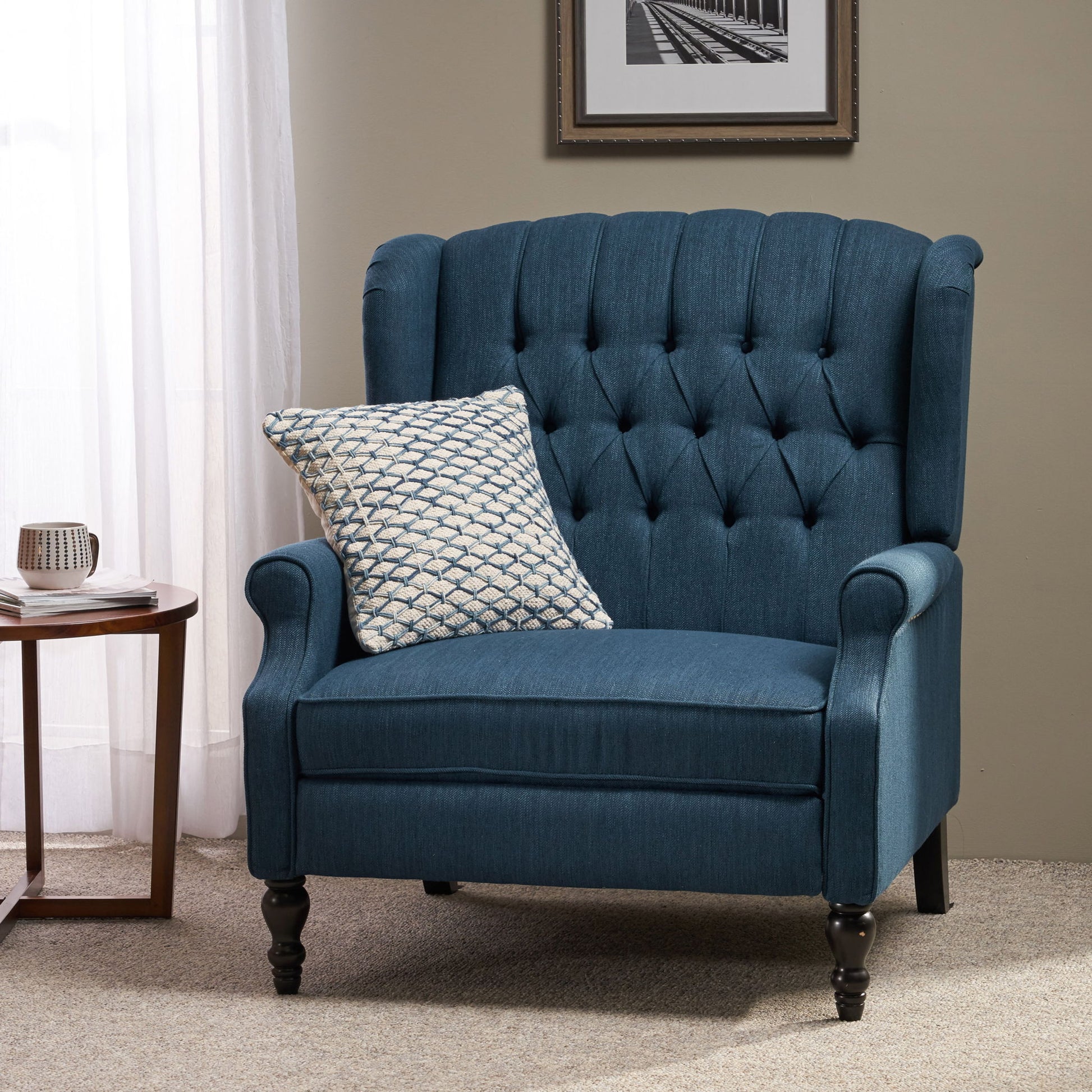 The Dunham Contemporary Fabric Tufted Oversized Pushback Recliner with curved armrests and dark wooden legs sits on a beige carpet, paired with a patterned cushion. Nearby is a round wooden table with a mug, and a framed photo hangs on the wall.