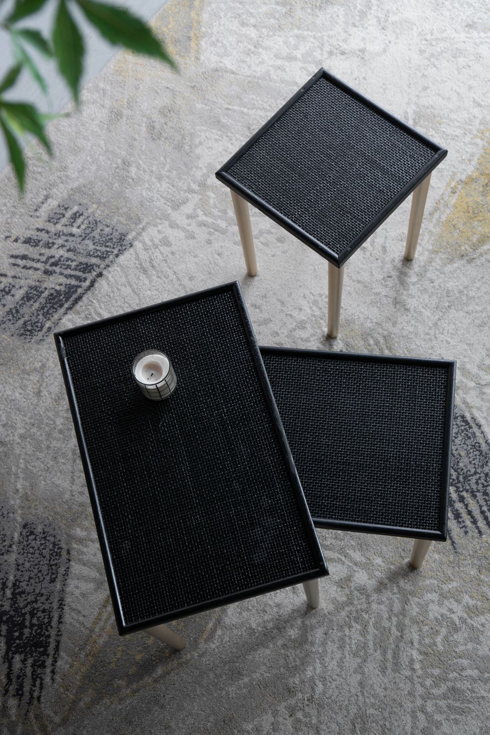 Three black side tables with gold legs are arranged on a patterned gray rug. One rectangular table holds a small candle, and a green plant sits in the top left corner. Product: Side Tables (Set of 3) - Black / Gold.