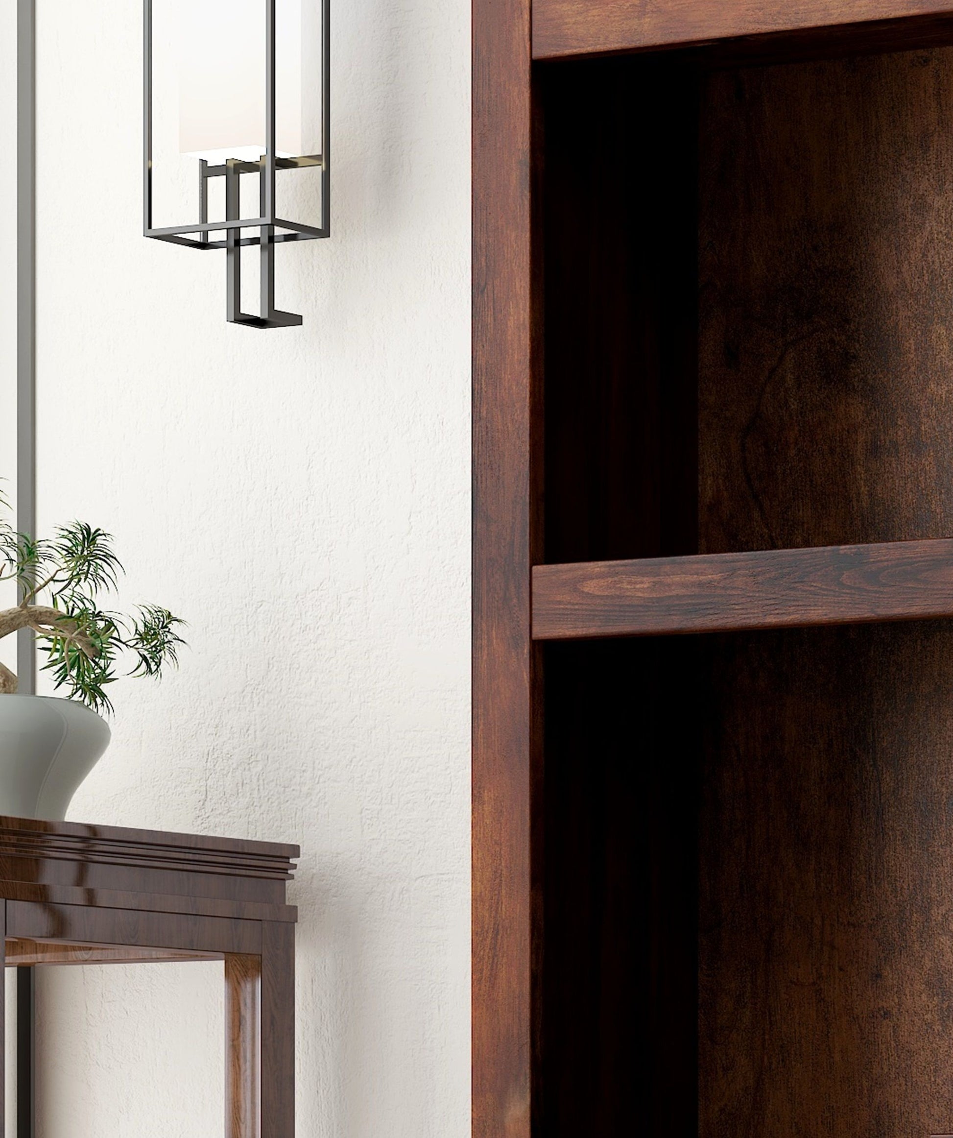 A close-up of the Sausalito - High Bookcase beside a wooden table with a potted plant. A modern wall-mounted light fixture hangs above against a white textured wall.