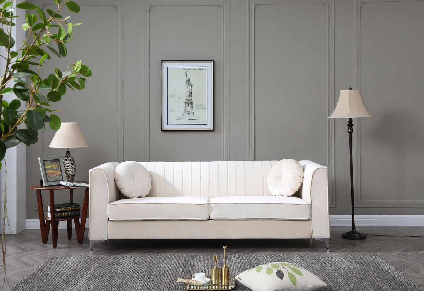 A modern living room featuring the Delray Micro Suede Sofa in beige, two round pillows, a gray wall, framed artwork, two lamps, a side table with books, a potted plant, and a gray rug with a decorative pillow and tray on the floor.