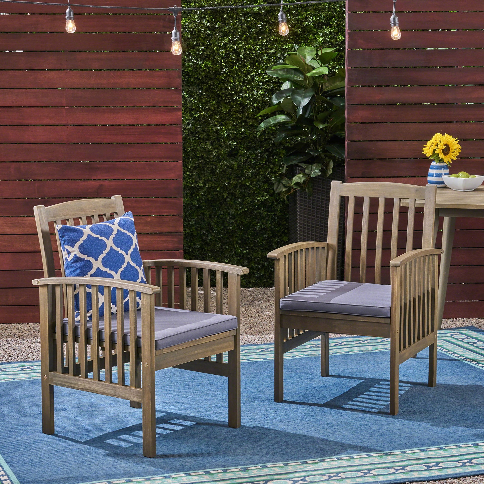 Two Casa - Acacia Dining Chairs with gray cushions rest on a blue rug. One has a blue patterned pillow. Behind them are red wood panels, green plants, string lights, and a small table topped with sunflowers.