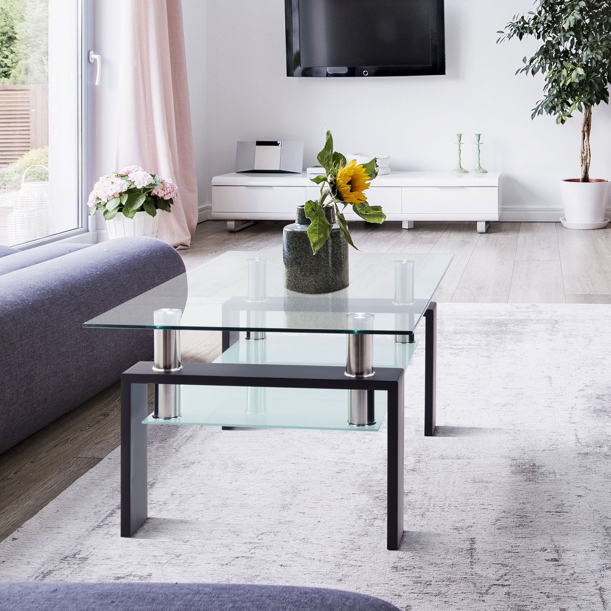 A modern living room featuring a Modern Rectangular Coffee Table With Tempered Glass Top holding a sunflower vase, light rug, gray sofas, wall-mounted TV, and potted plants by a sunlit window.