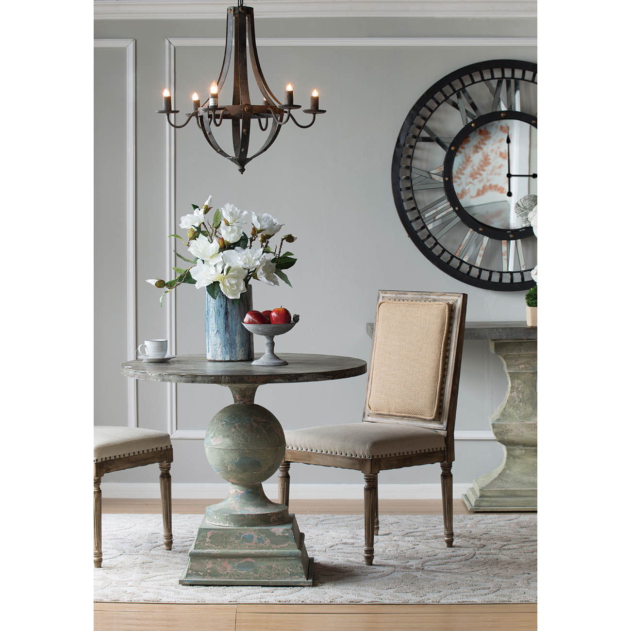 Elegant dining area with a round table, two upholstered chairs, a vase of white flowers, fruit bowl, large wall clock, and a 6 Light Metal Chandelier in black with adjustable chain (bulb not included), in a classic neutral-toned setting.