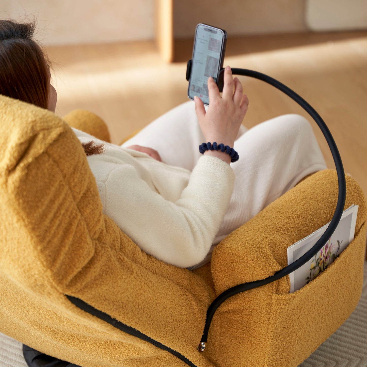 A person in light clothes sits on an Adjustable Swivel Lounge Chair with 360° rotating seat and headrest, watching their smartphone held by a flexible holder. A magazine is tucked into the chair’s side pocket.