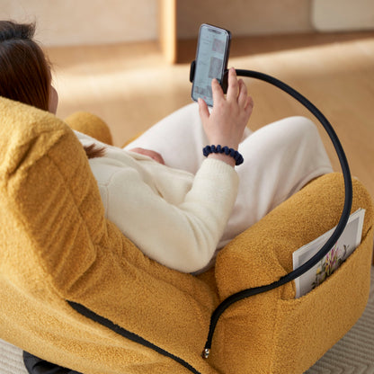 A person in light clothes sits on an Adjustable Swivel Lounge Chair with 360° rotating seat and headrest, watching their smartphone held by a flexible holder. A magazine is tucked into the chair’s side pocket.