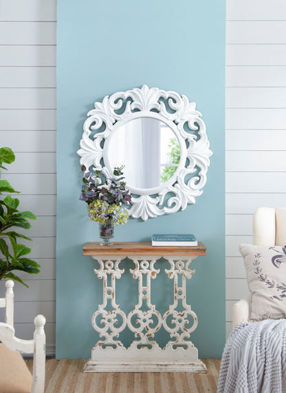 A decorative white mirror hangs on a light blue wall above the Classic Vintage Wash Console Table with Wood Top in Brown/Antique White, which holds a vase of flowers and a book. A nearby plant and part of a couch complete the scene.