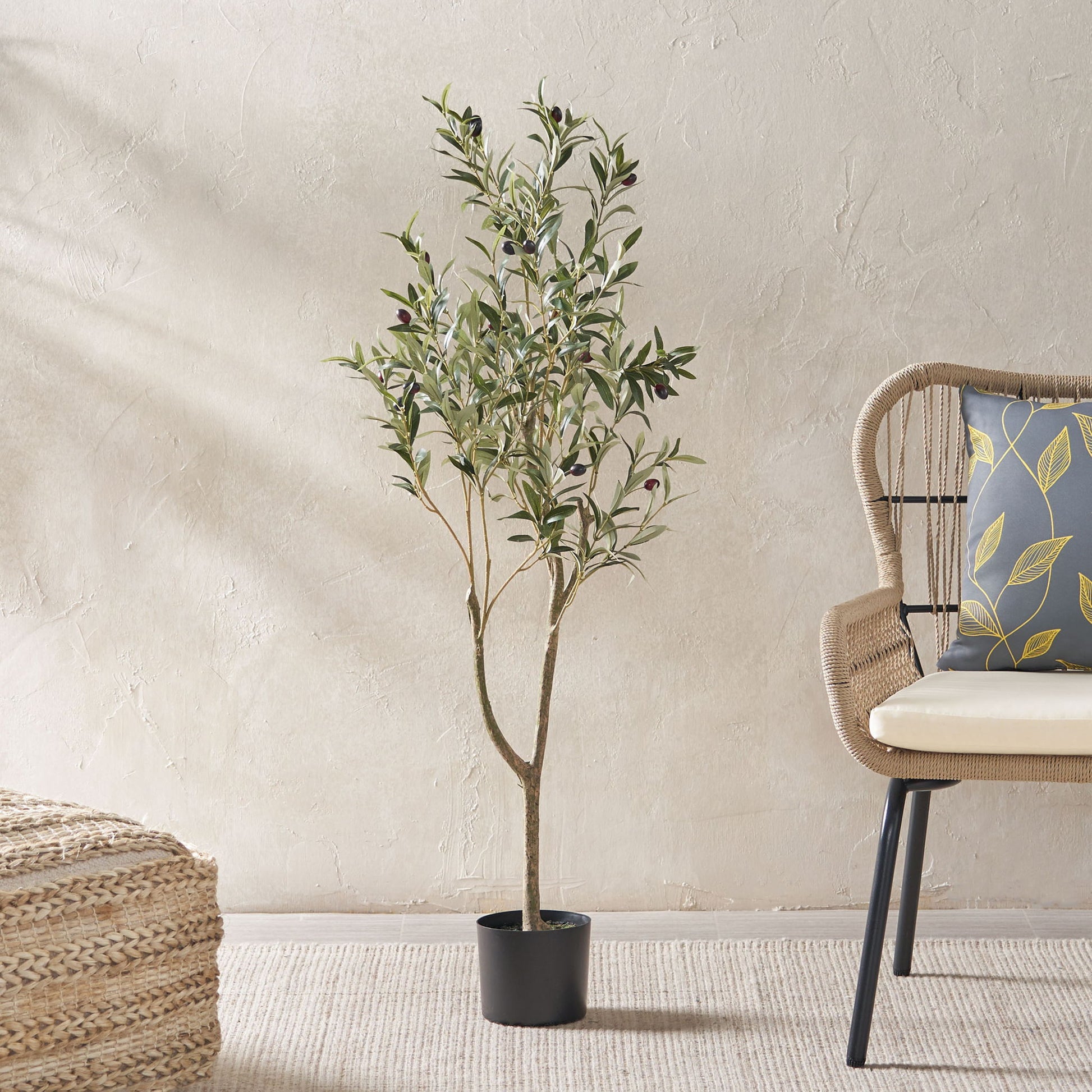 An Artificial Olive Tree with slender branches and green leaves is placed beside a wicker chair with a patterned cushion in a minimalist room featuring a textured beige wall and woven rug.