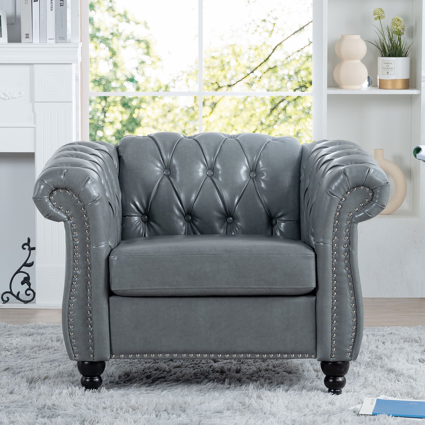 A 1 Seater Sofa For Living Room, featuring gray tufted leather upholstery with rolled arms and nailhead trim, sits on a light gray rug in a bright living room with a large window and shelves in the background.