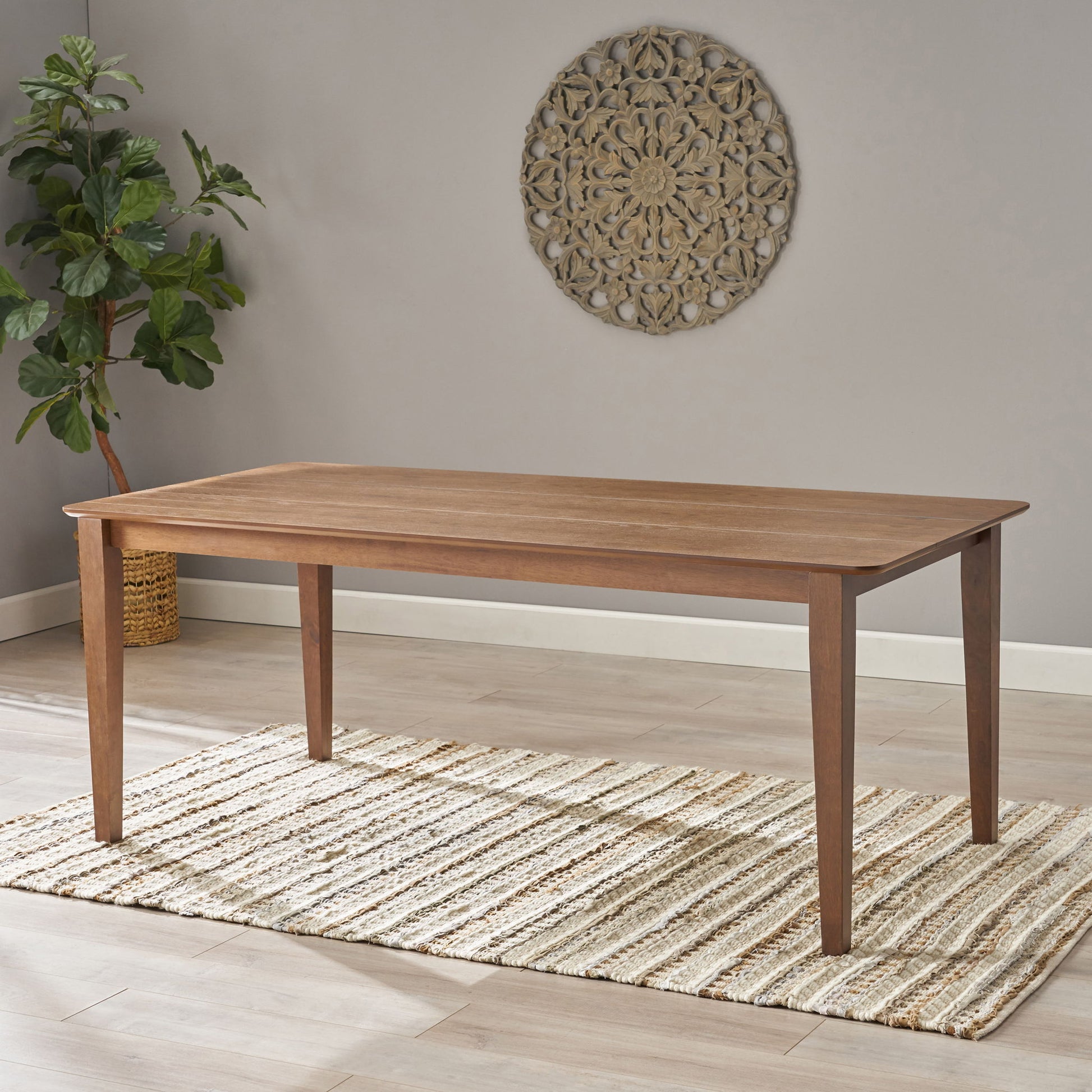 The Farmhouse Charm Dining Table With Square Leg sits on a striped rug in a bright room with light wood floors, a potted plant in the corner, and a round wall hanging on the gray wall.
