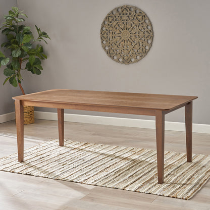 The Farmhouse Charm Dining Table With Square Leg sits on a striped rug in a bright room with light wood floors, a potted plant in the corner, and a round wall hanging on the gray wall.
