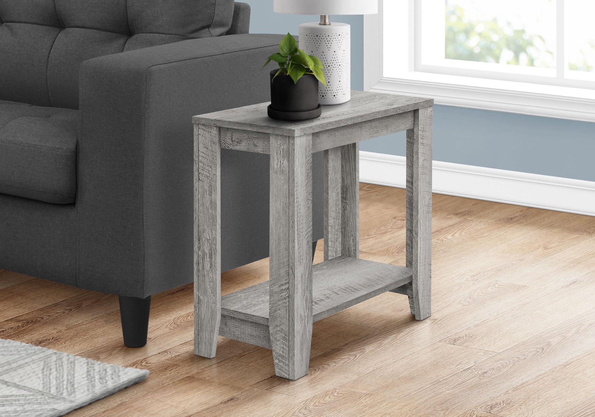 An Accent Table, Side Transitional with an elegant design, in gray wood, featuring a shelf and topped with a potted plant and white lamp, stands next to a dark gray sofa on light wood flooring by a window.