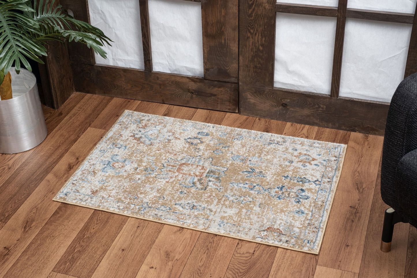 The Noble - GC_GEN7008 Area Rug, featuring muted blue, beige, and brown patterns, lies on a wooden floor in front of dark wooden doors, beside a potted plant and a black upholstered chair.