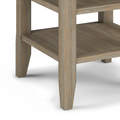 Close-up of the Acadian - Handcrafted End Table’s lower corner, featuring two shelves, a natural wood grain finish, and square tapered legs on a white surface.