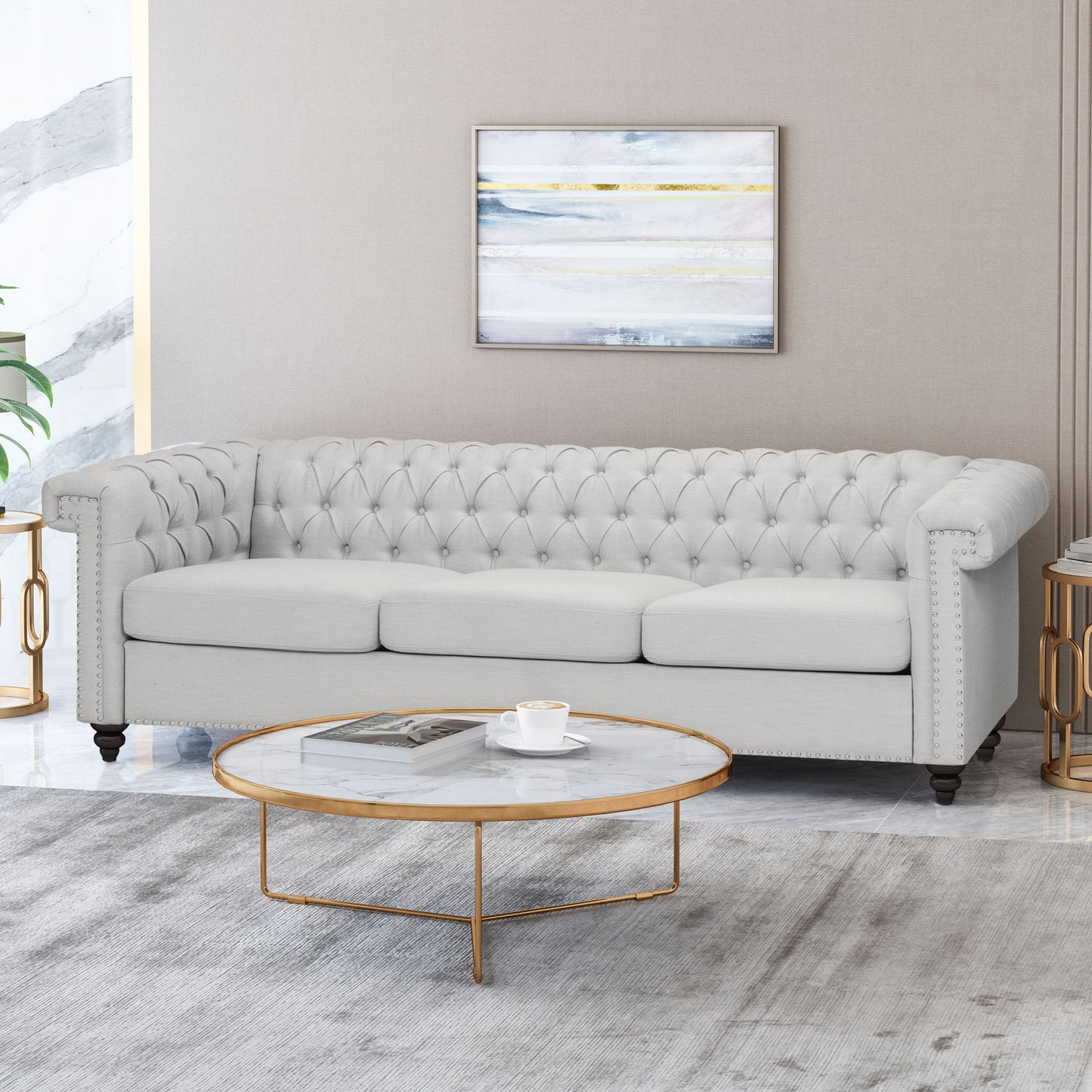 A modern living room with a Chesterfield Style 3 Seater Sofa With Deep Button Tufting in light gray, a round marble coffee table with gold legs, cup and saucer, side tables, abstract art on a beige wall, and a light gray rug.