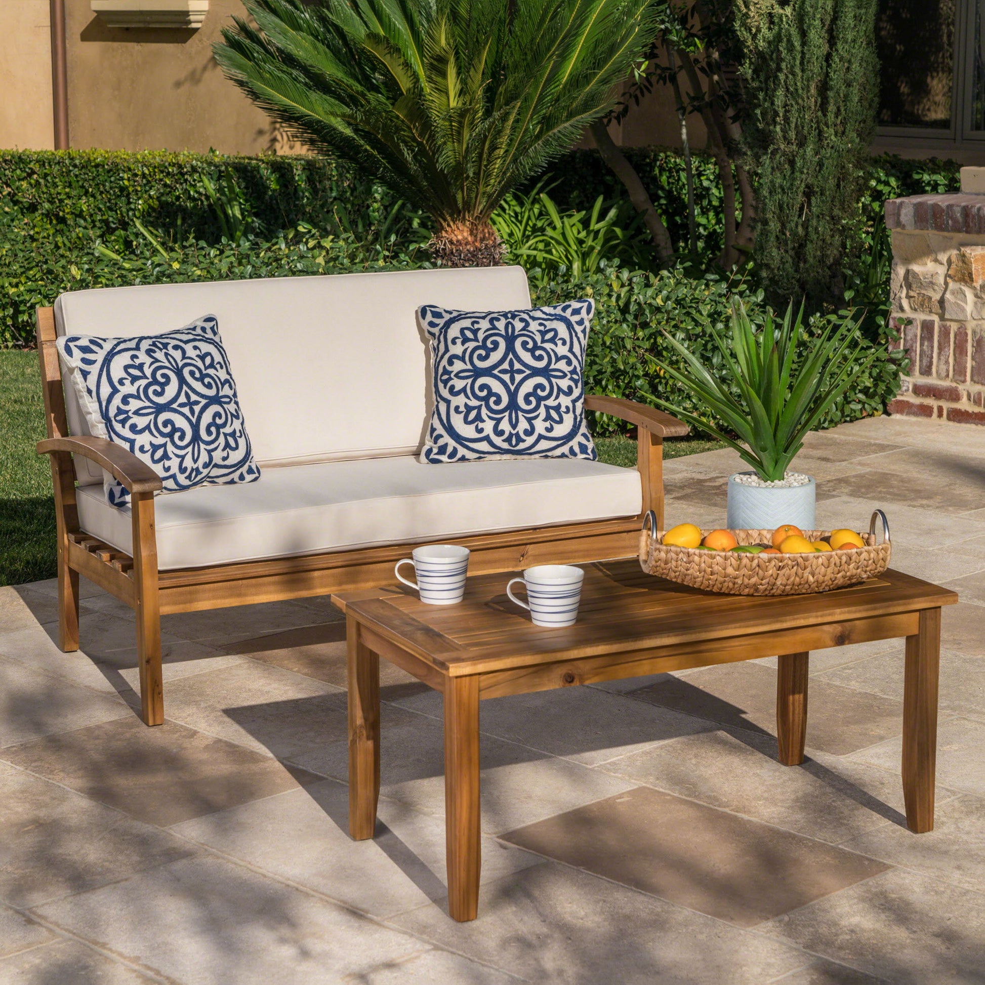 The Outdoor Chat Set With Coffee Table And Loveseat features cream cushions, two blue and white pillows, and sits by a matching table with mugs and a basket of oranges, surrounded by lush greenery.
