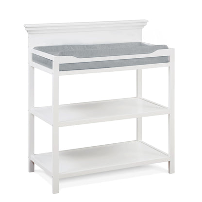 The Universal - Changing Table features a white wooden frame, two open shelves for storage, and a grey cushioned changing pad on top.
