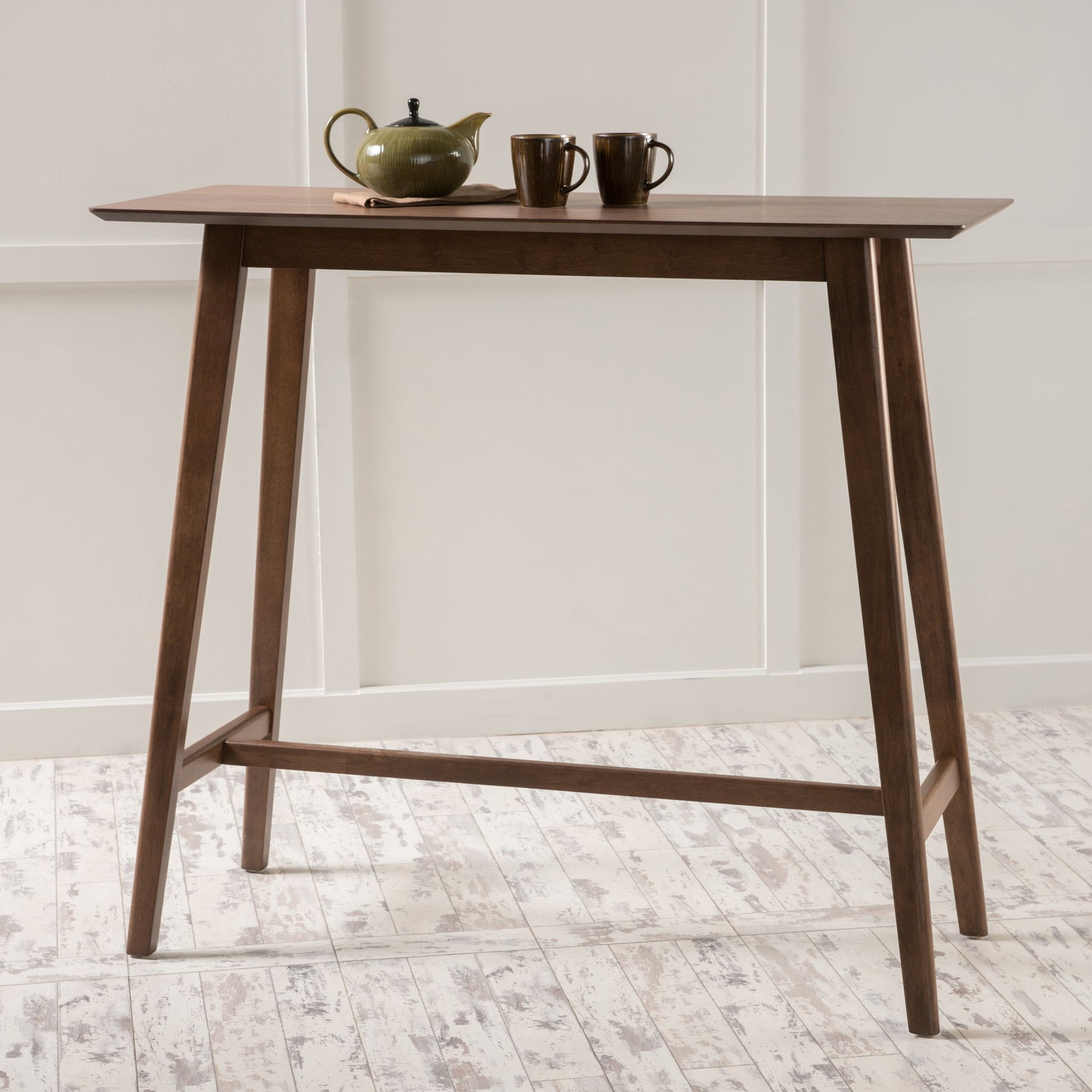 A Bar Height Dining Table With Splayed Legs And Elongated Rectangular Top stands on a white textured floor. On the tabletop are a green teapot with a lid and two matching ceramic cups. White paneled walls form the background.